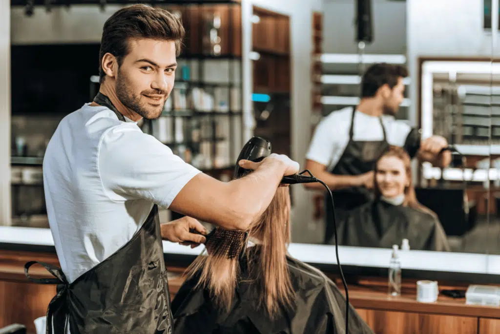 young hairstylist smiling at camera while drying hair to woman in beauty salon